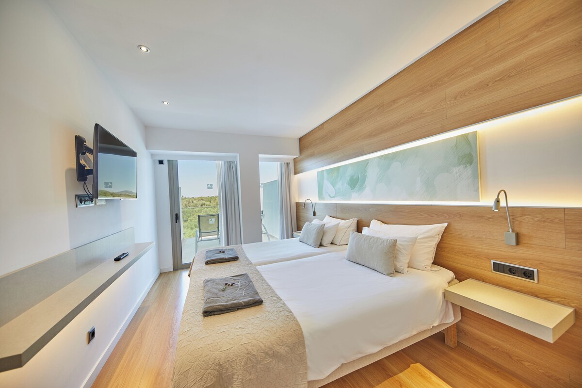 A modern hotel room with two beds, a large window, a wooden headboard, and a built-in bench.