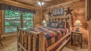 Ein Schlafzimmer einer Holzhütte mit einem großen Bett, einer Sterndeko, einer Lampe und einem Fenster mit Blick auf Bäume.