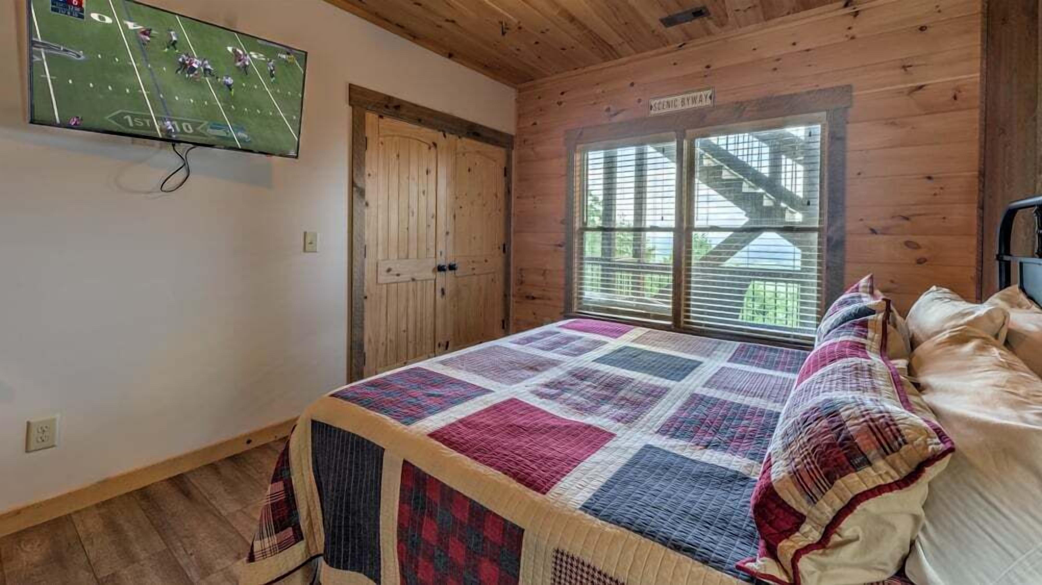 A bedroom with a large bed, a television mounted on the wall, a wooden door, and a window with blinds.