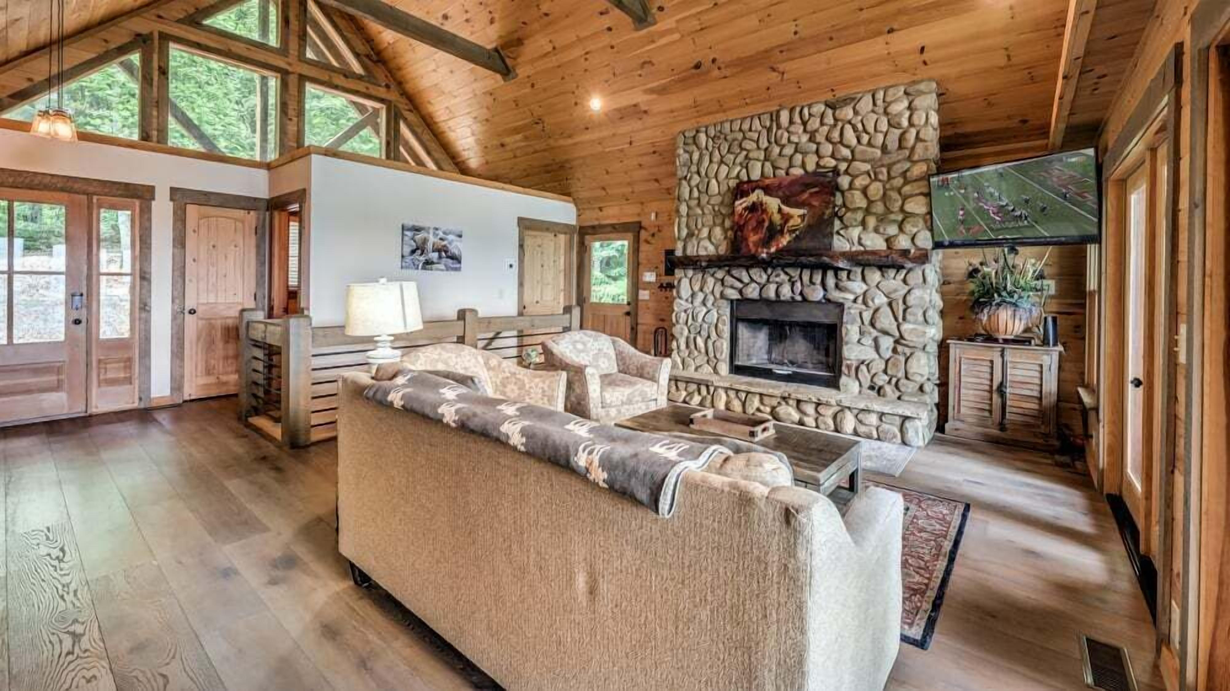 A cozy cabin interior with a stone fireplace, wooden beams, and a large window overlooking the forest.