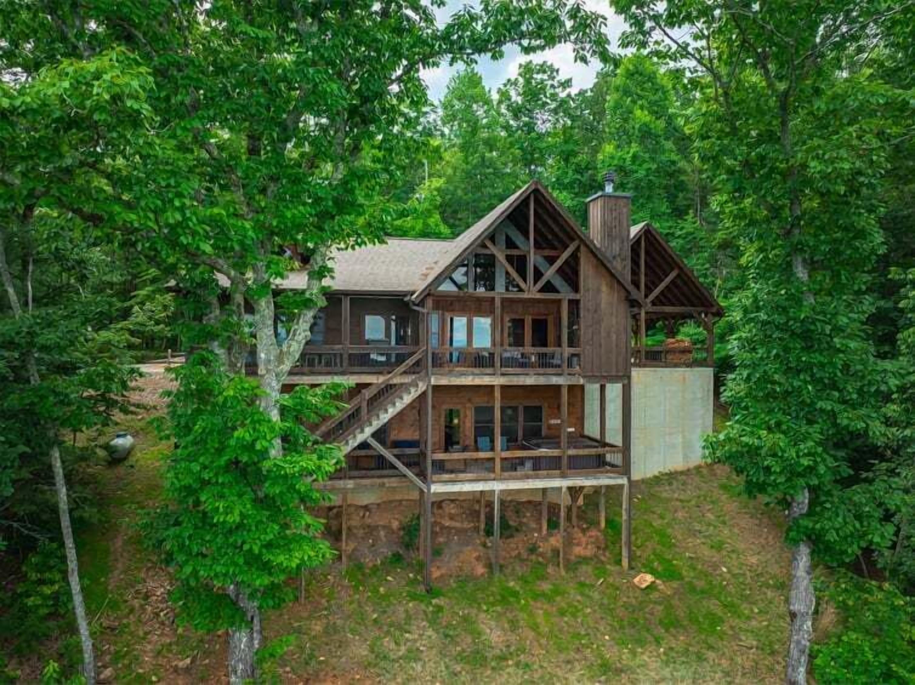 Une cabane en bois à deux étages, dotée d’un balcon et d’une cheminée, entourée d’une forêt dense.