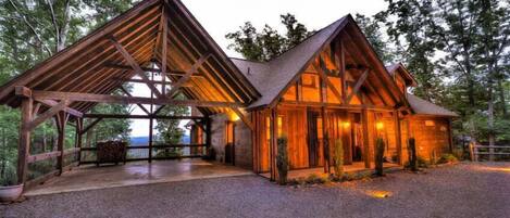 A rustic cabin with a large open porch and a covered area, surrounded by trees.