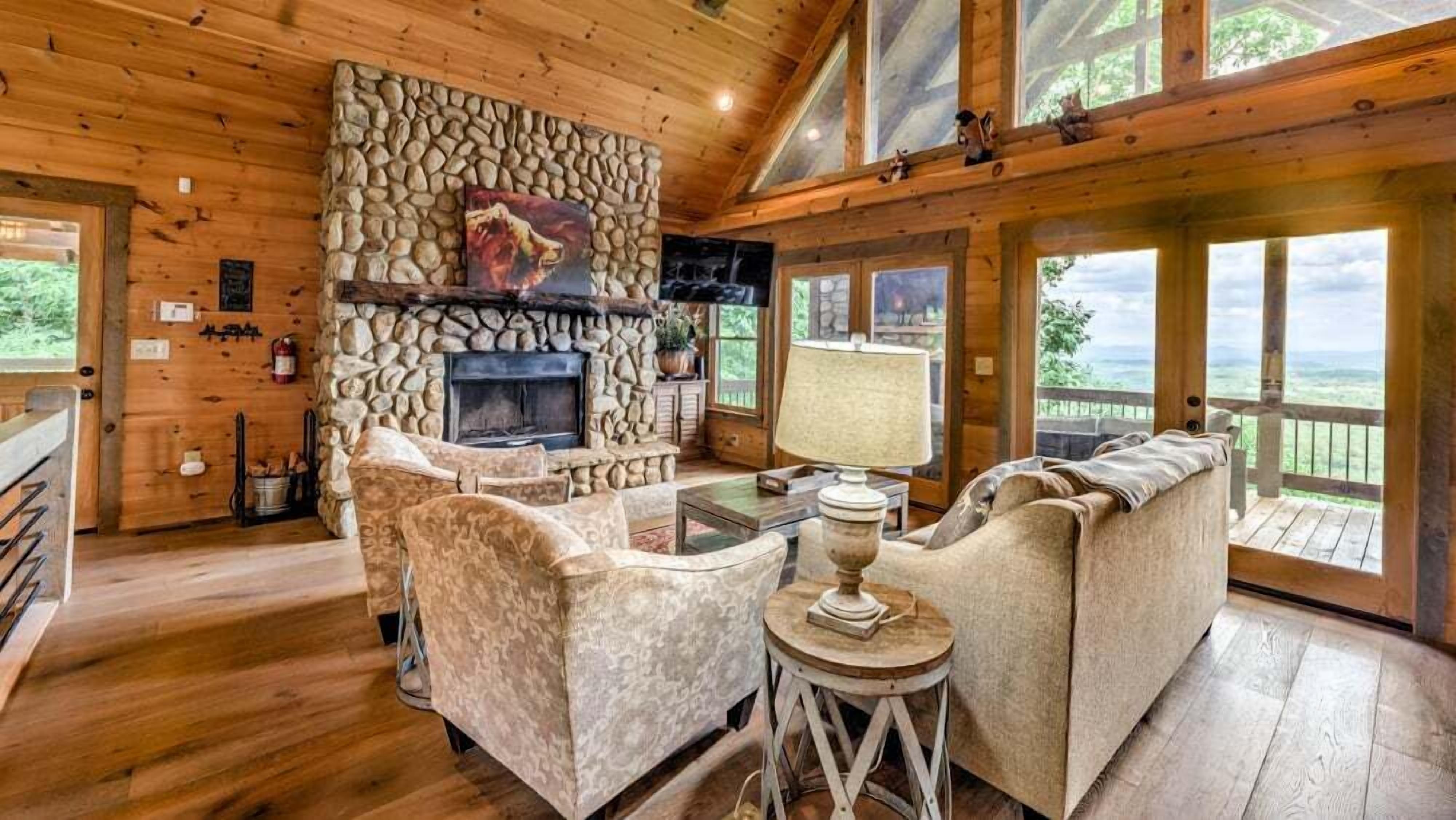 A cozy cabin interior with a stone fireplace, comfortable seating, and large windows offering scenic views.