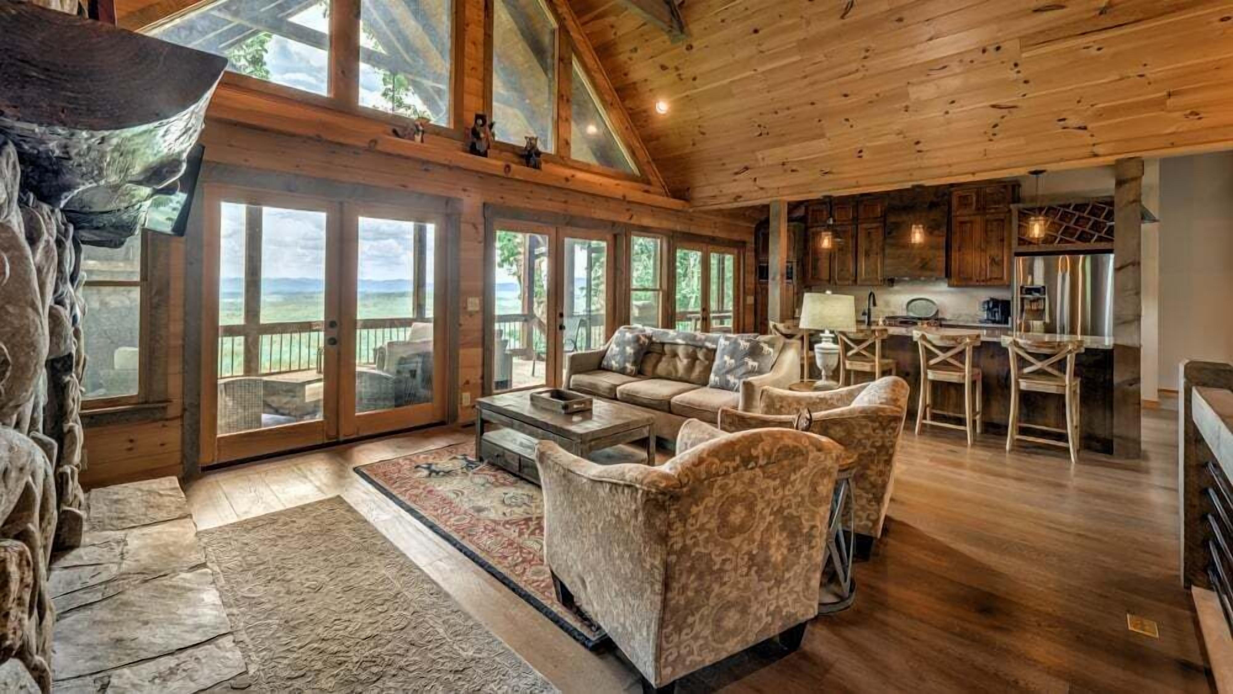 A spacious living room with a stone fireplace, large windows, and a kitchen area with wooden cabinets.