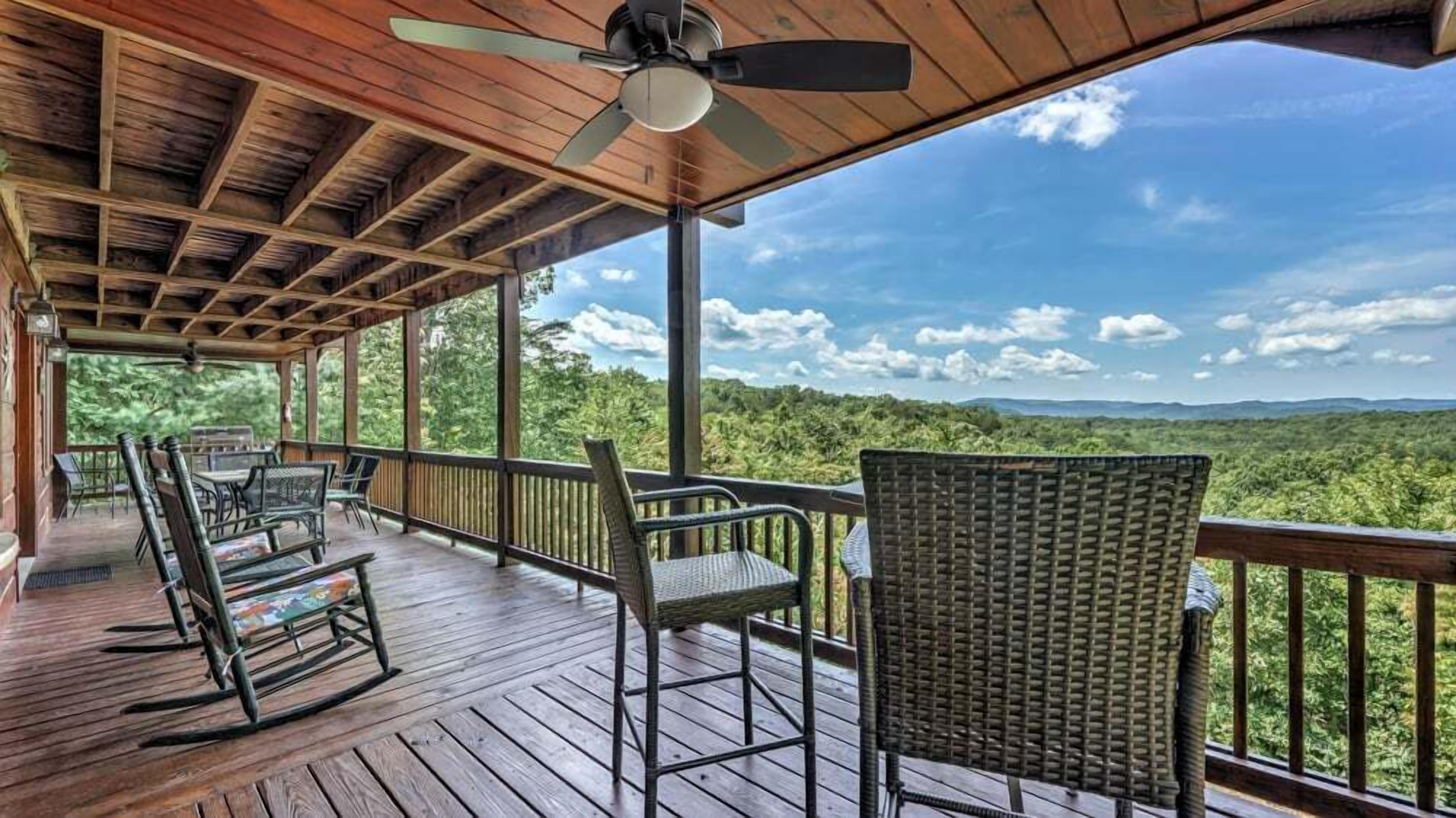 A wooden deck with outdoor furniture, a ceiling fan, and a view of a forested landscape.