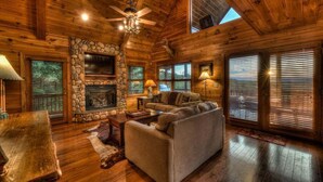 A log cabin interior with a stone fireplace, a ceiling fan, and a large window with blinds. - Big Sky Retreat- Pet Friendly | Game Room | Hot Tub with Views (Blue Ridge)