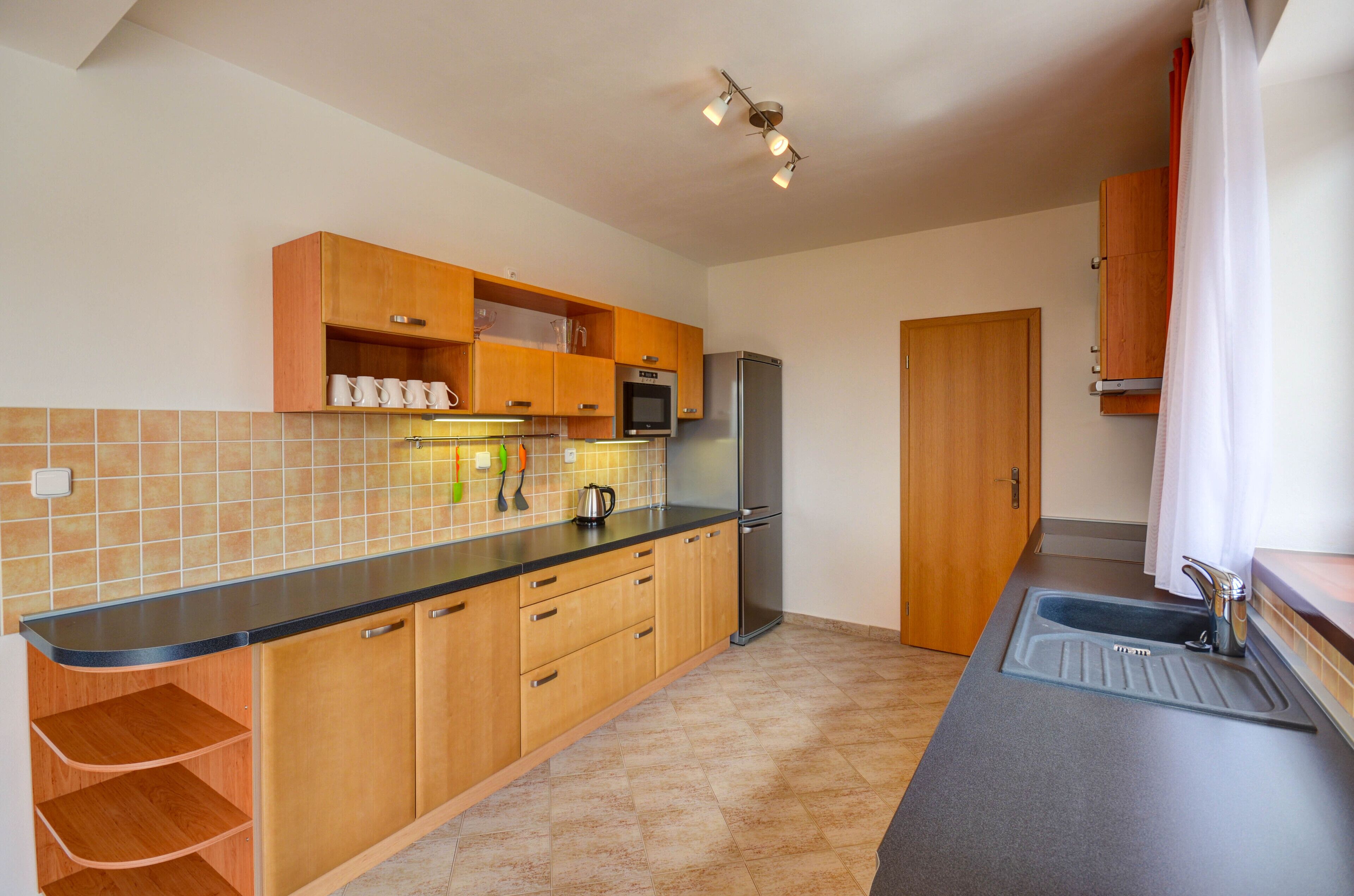 A kitchen with wooden cabinets, a black countertop, a stainless steel refrigerator, and a sink.
