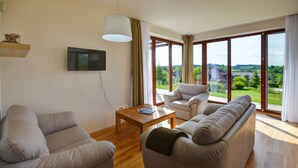 A modern living room with a sofa, armchair, and coffee table. A flat-screen TV is mounted on the wall. Large windows offer a view of a green landscape.