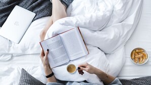 A person lying on a bed with a laptop, a book, and a cup of coffee.
