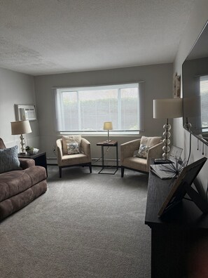 A living room with a brown sofa, two armchairs, a coffee table, a TV, and a large window with blinds. - Cozy 2 bedroom retreat on quiet Ledgewood Dr! Minutes to Downtown Portsmouth! (Portsmouth)
