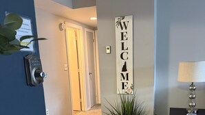 A hallway with a welcome sign, a potted plant, a lamp on a table, and a door with a lock.