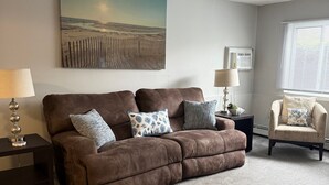 A living room with a brown sofa, two side tables with lamps, a chair, a painting on the wall, and a window with blinds.