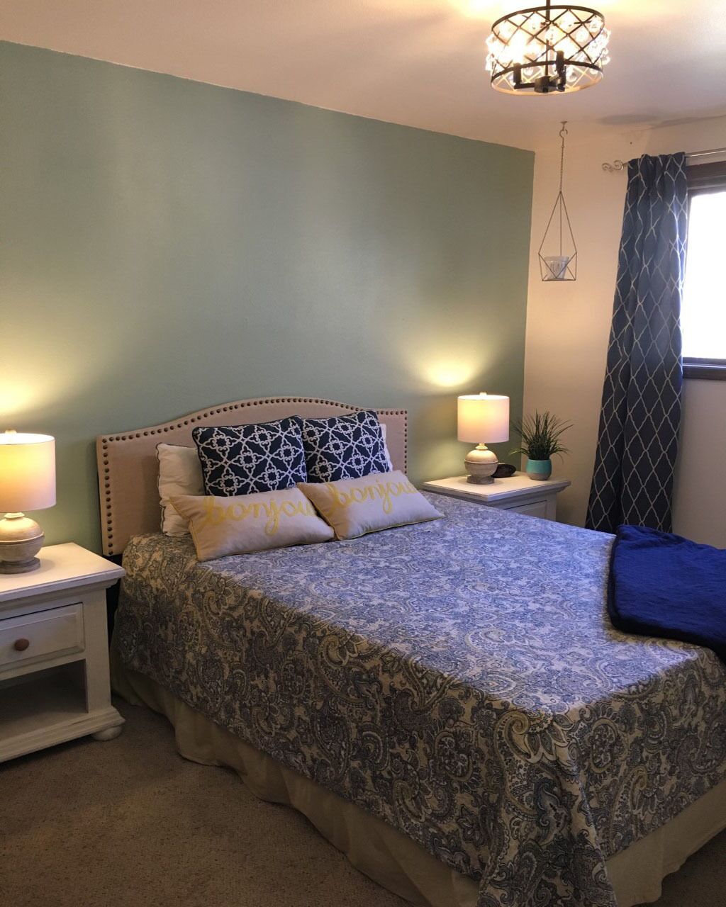 A bedroom with a bed, two bedside tables, a nightstand, a lamp, a potted plant, and a window with curtains.