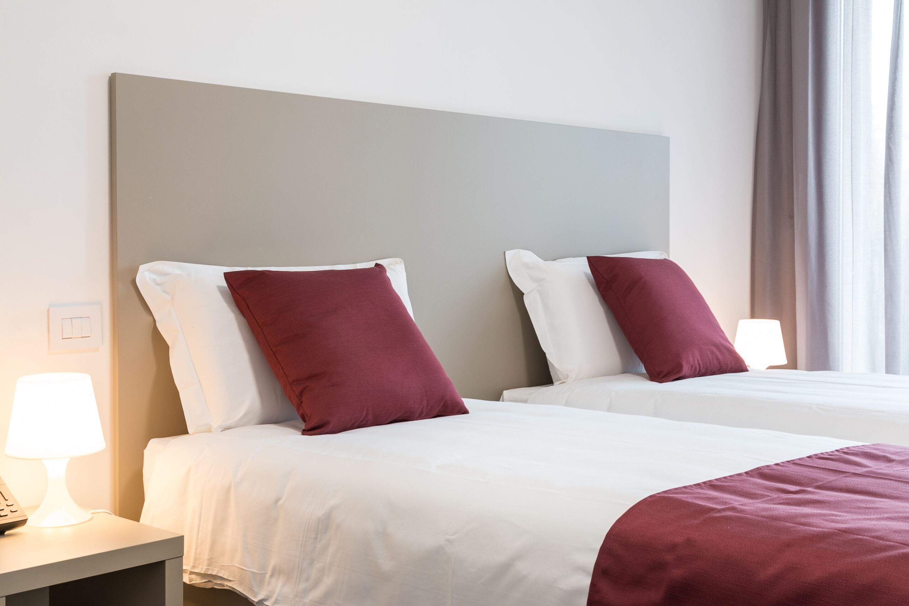 A hotel room with two beds, white bedding, and red pillows. There is a nightstand with a lamp and a phone.
