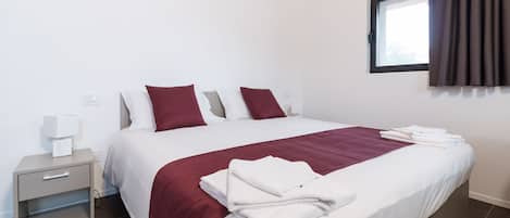 Una cama bien hecha con ropa de cama blanca y granate, una mesita de noche con lámpara y una ventana con cortinas.