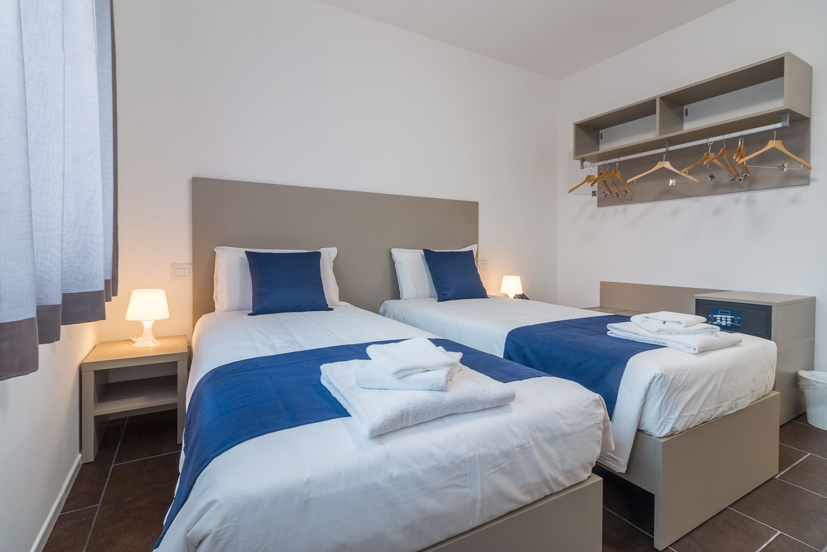 Habitación de hotel con dos camas, una mesita de noche con lámpara, un armario con perchas y un pequeño cubo de basura.