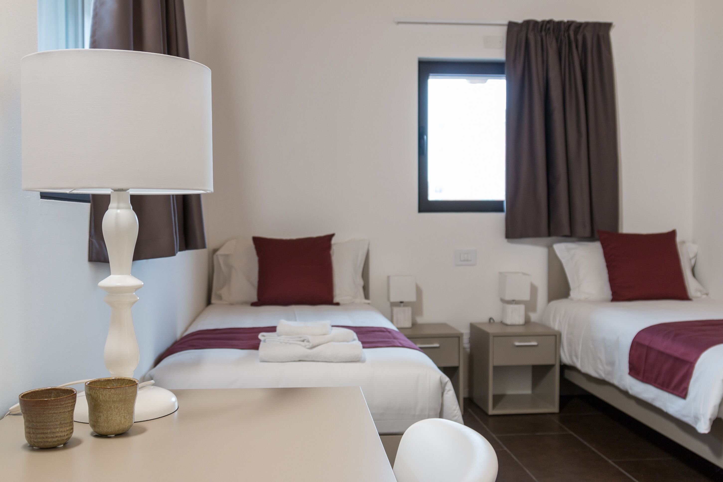 Een hotelkamer met twee bedden, een tafel met een lamp, twee nachtkastjes en een raam met gordijnen.