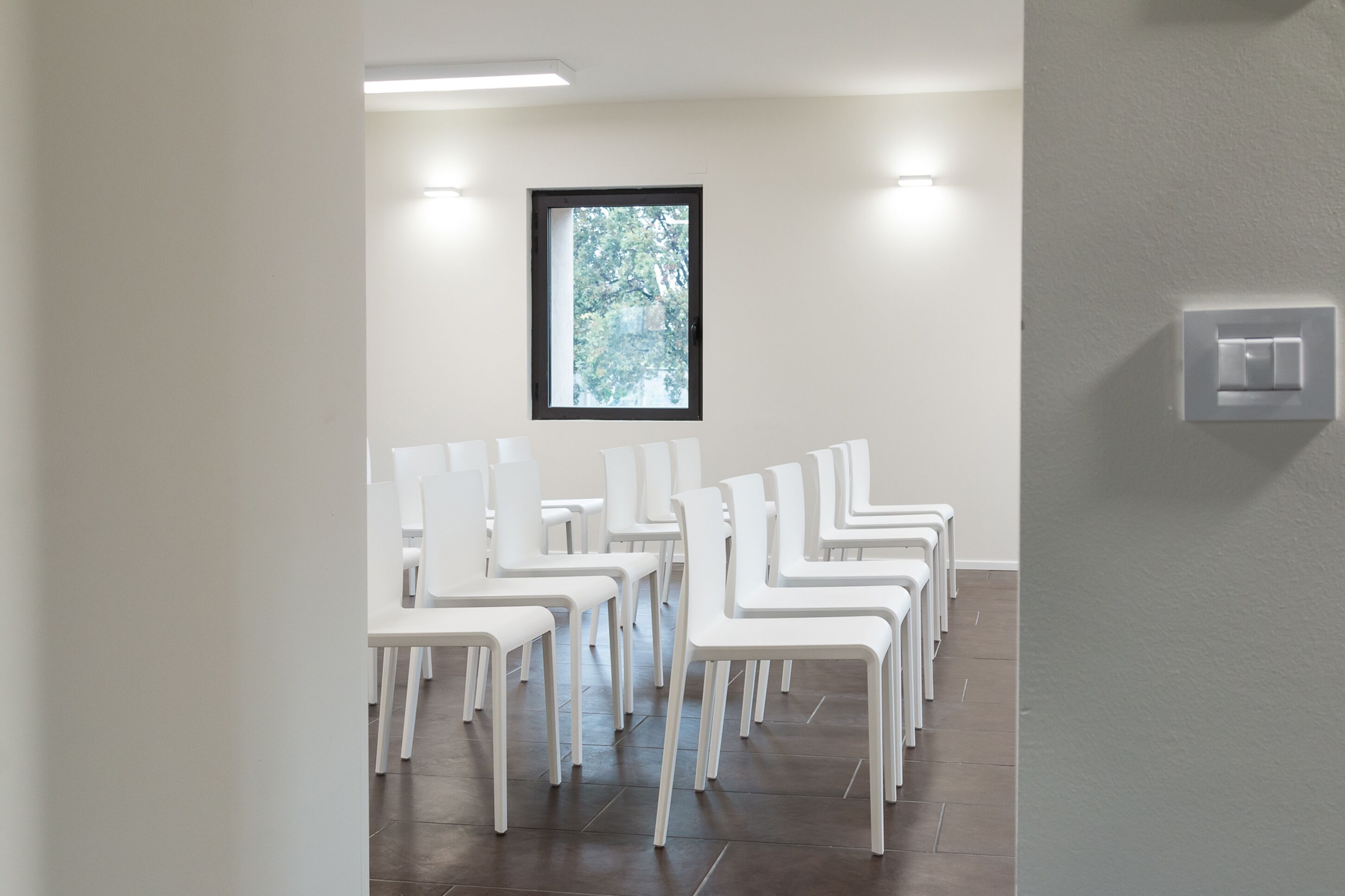 A room with white chairs arranged in rows, a window with a view of trees, and a light switch on the wall.