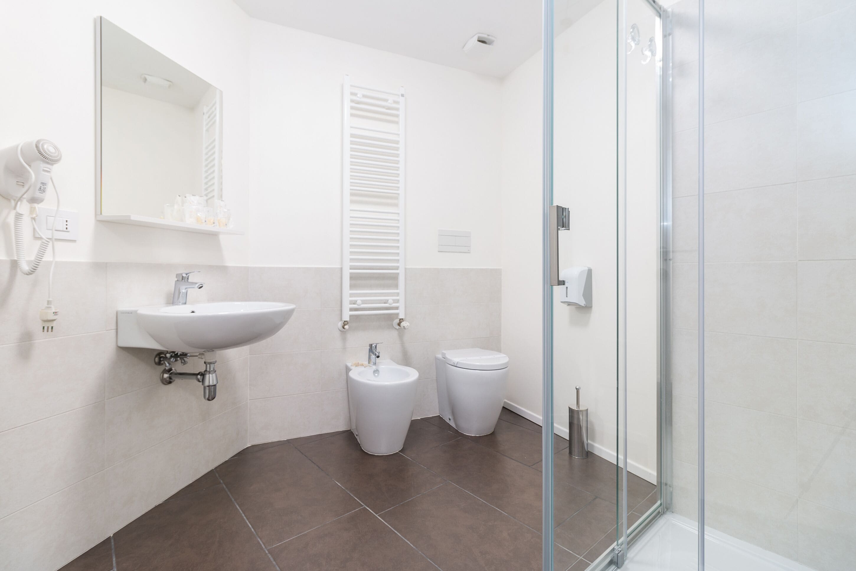 Un baño moderno con un lavamanos blanco, un bidé y un inodoro, un box de ducha de vidrio y un secador de pelo en la pared.