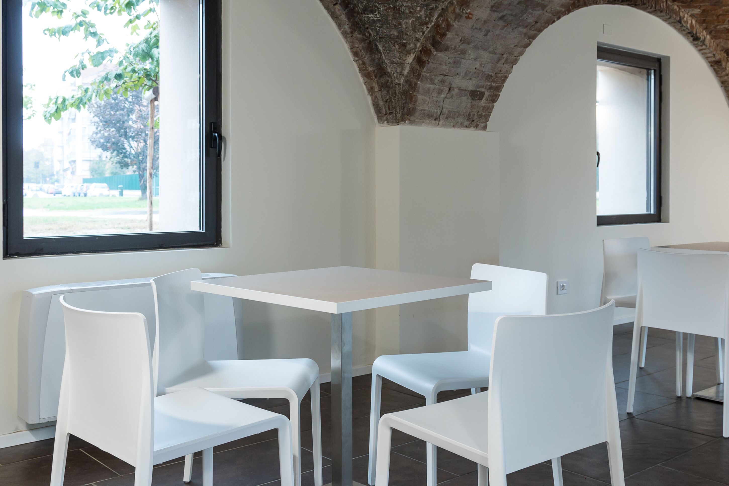 A dining area with white tables and chairs, a brick ceiling, and large windows with views of greenery.