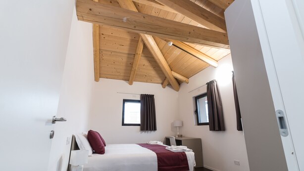 A bedroom with a wooden ceiling, a bed with a white and maroon bedspread, a bedside table with a lamp, and two windows with curtains. - Hotel Cascina Fossata & Residence (Turin)
