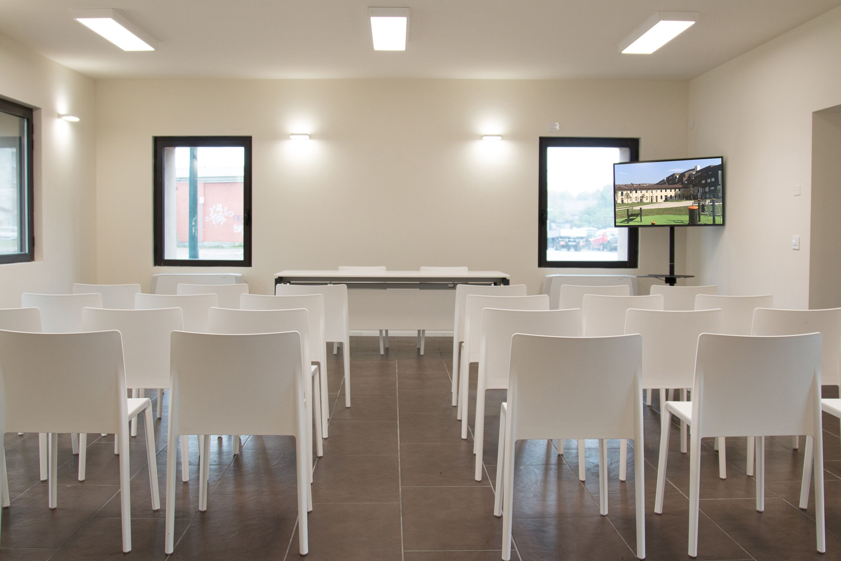 A conference room with white chairs arranged in rows, a large screen on the wall, and a window with a view of buildings.