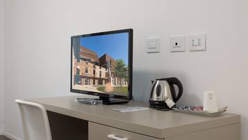 A hotel room with a desk, a monitor displaying a building, a kettle, and a tray with items.