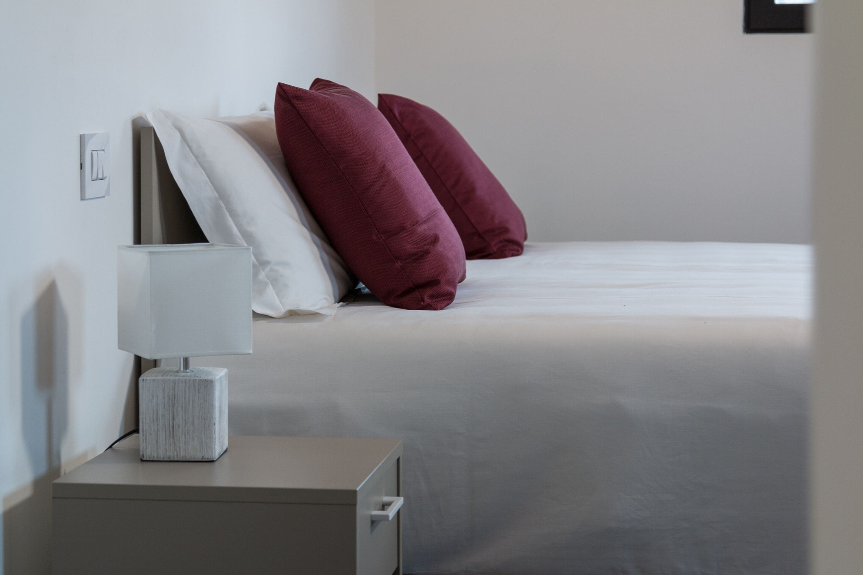 A neatly made bed with white and maroon pillows, a bedside table with a lamp, and a wall-mounted light switch.