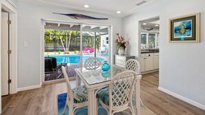 Zona pranzo con tavolo in vetro e sedie in vimini, cucina con mobili bianchi e vista su una piscina all'aperto.