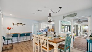 Un'area pranzo con tavolo in legno, sedie bianche e una decorazione a forma di pesce appesa. Si intravede una cucina sullo sfondo, con un forno a microonde e uno schienale piastrellato blu.