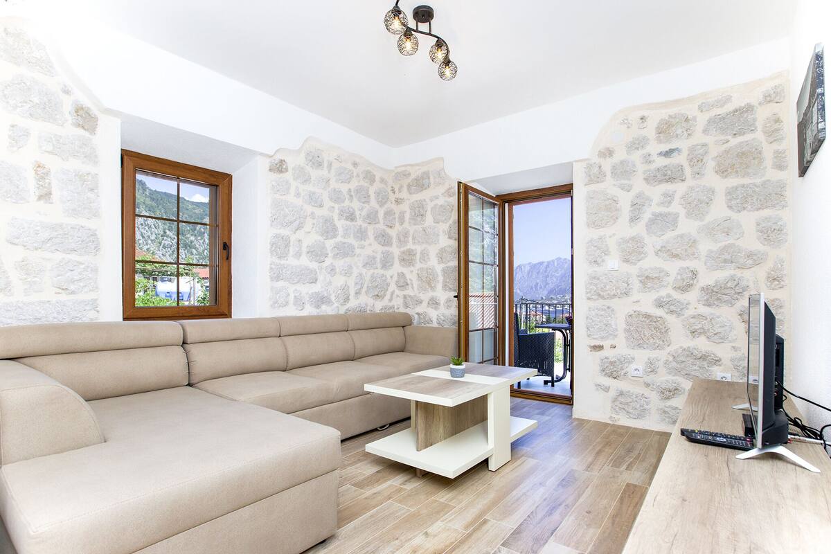A modern living room with a beige sectional sofa, a wooden coffee table, and a flat-screen TV on a wooden stand. The room features stone walls and large windows.