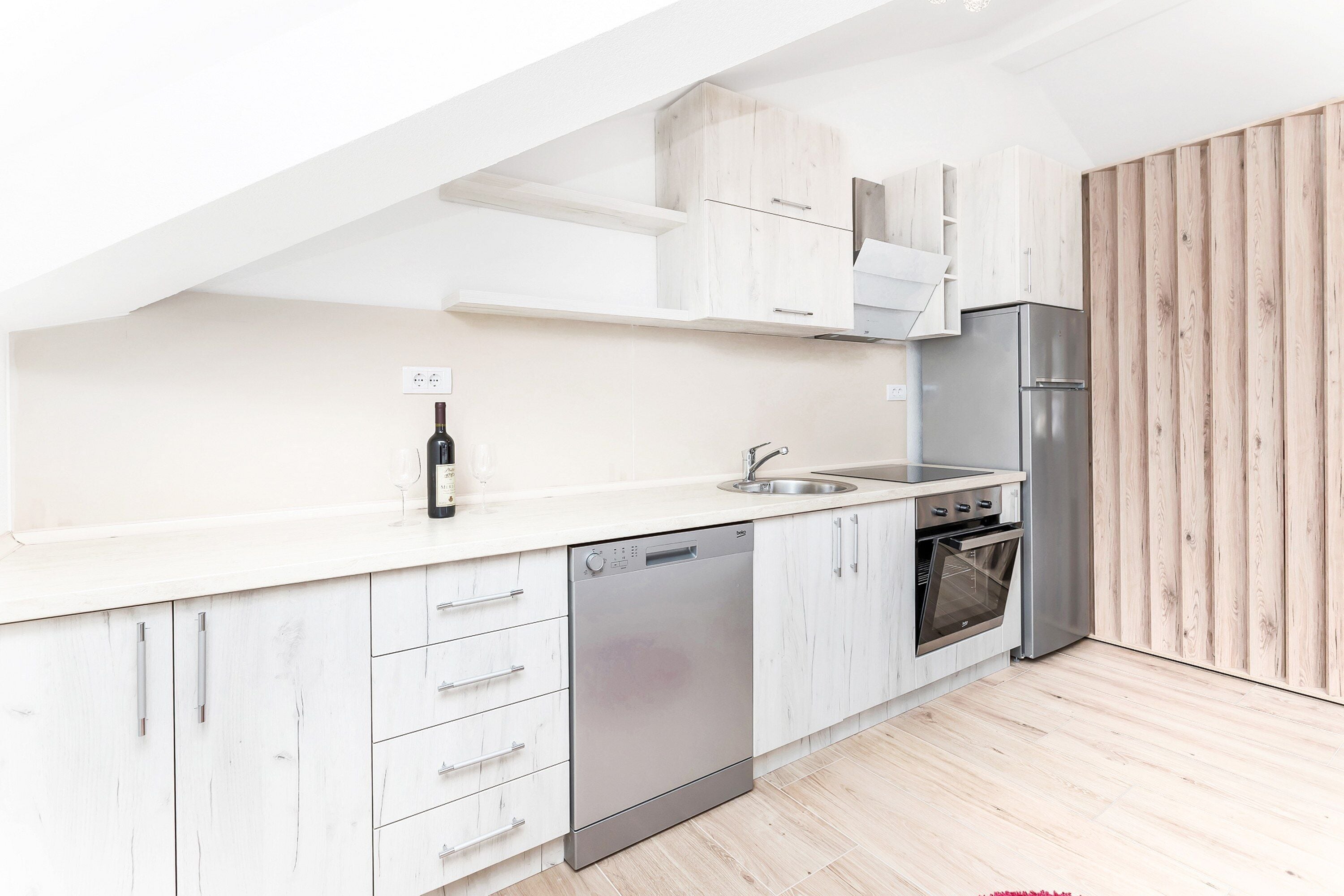 A modern kitchen with white cabinetry, a dishwasher, a stainless steel refrigerator, and a wooden accent wall.