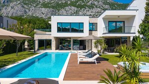 A modern two-story house with a swimming pool, outdoor seating, and a mountain backdrop.