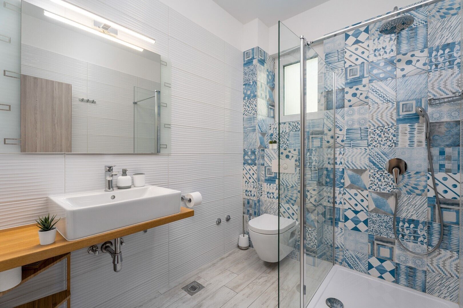Ein modernes Badezimmer mit einer gläsernen Duschkabine, einem weißen Waschbecken mit Holzablage und Wandfliesen mit Muster.