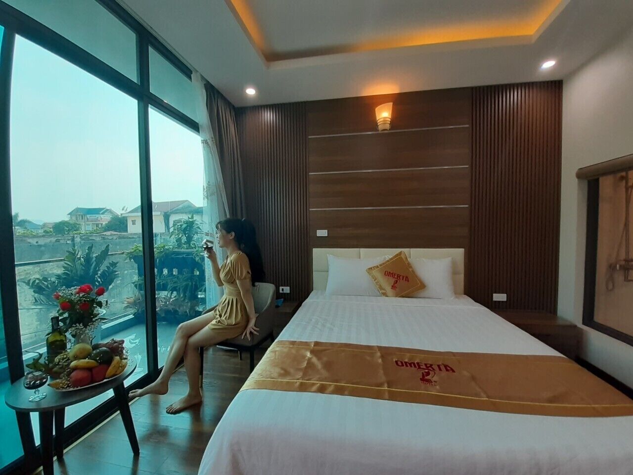 Habitación de hotel con una cama grande, una mesita de noche con un frutero y vistas a la ciudad a través de amplios ventanales.