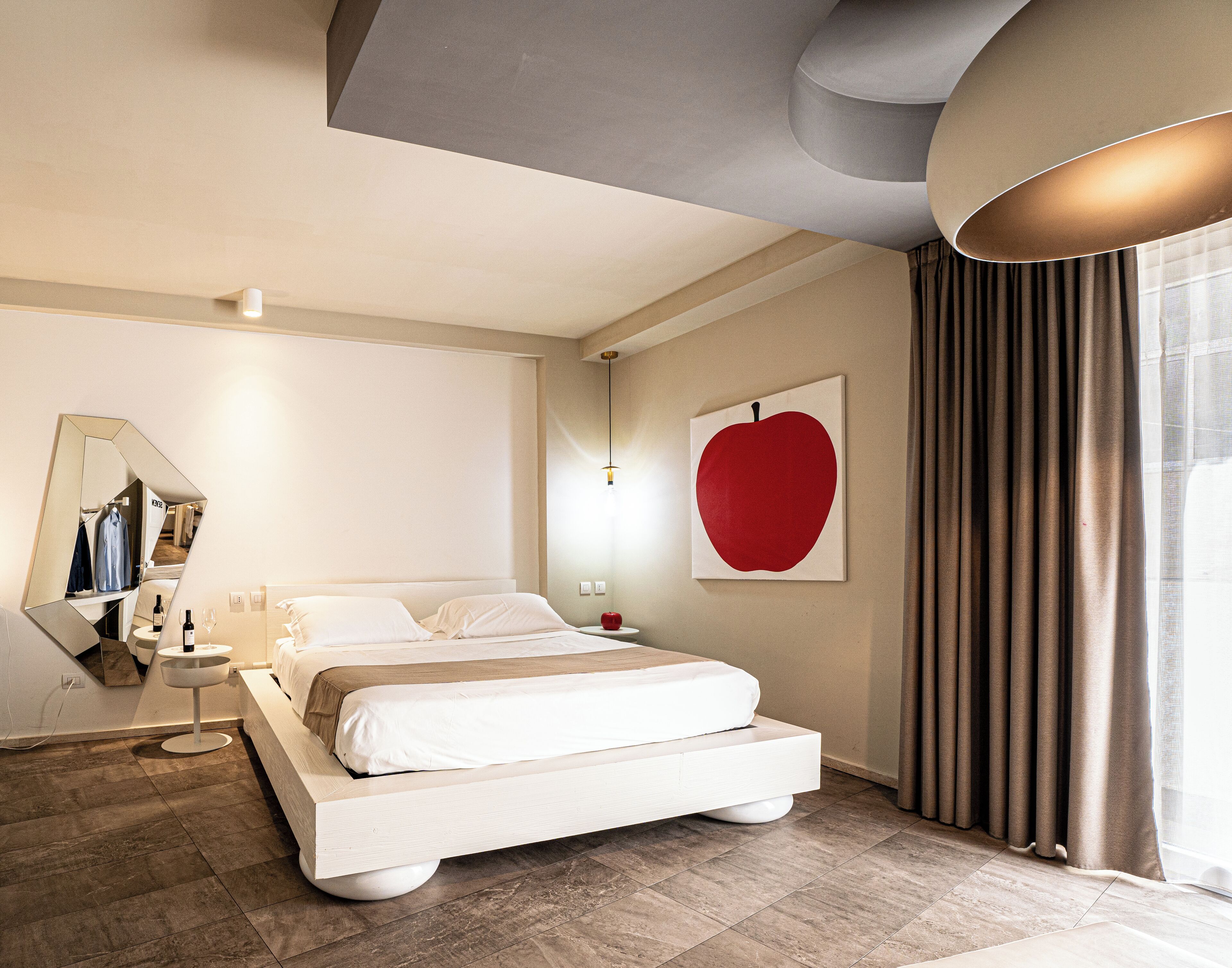 A modern hotel room with a large bed, a bedside table, a mirror, and a red apple artwork on the wall.