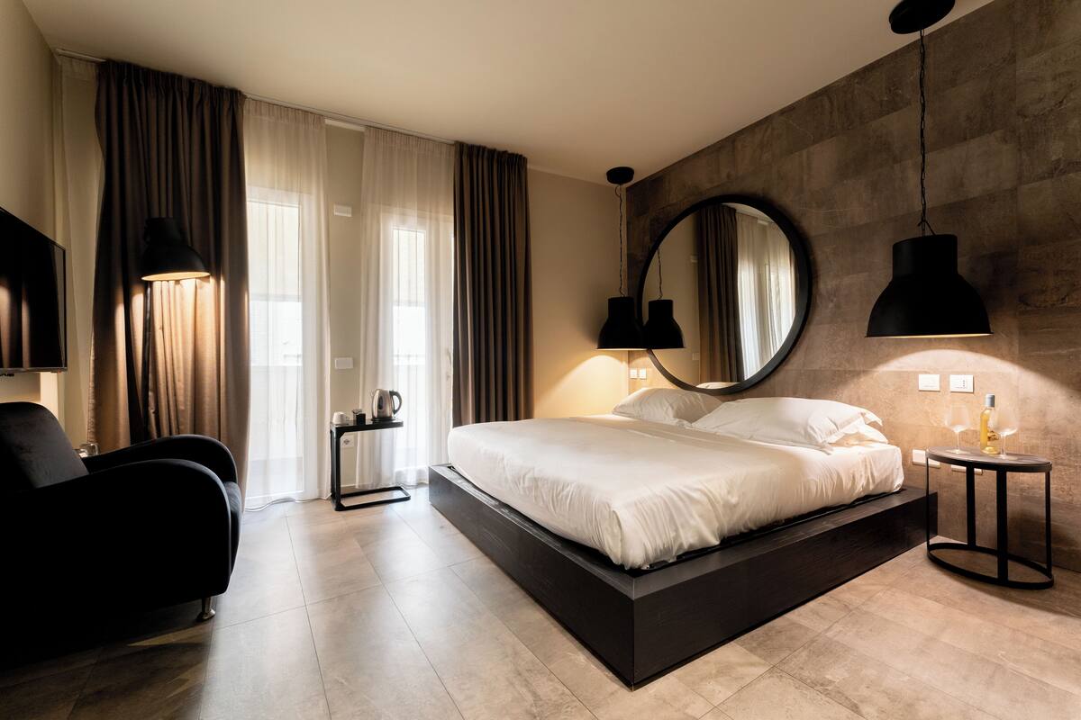 A modern hotel room with a large bed, a black armchair, a round mirror, and a small table with a bottle and glasses.