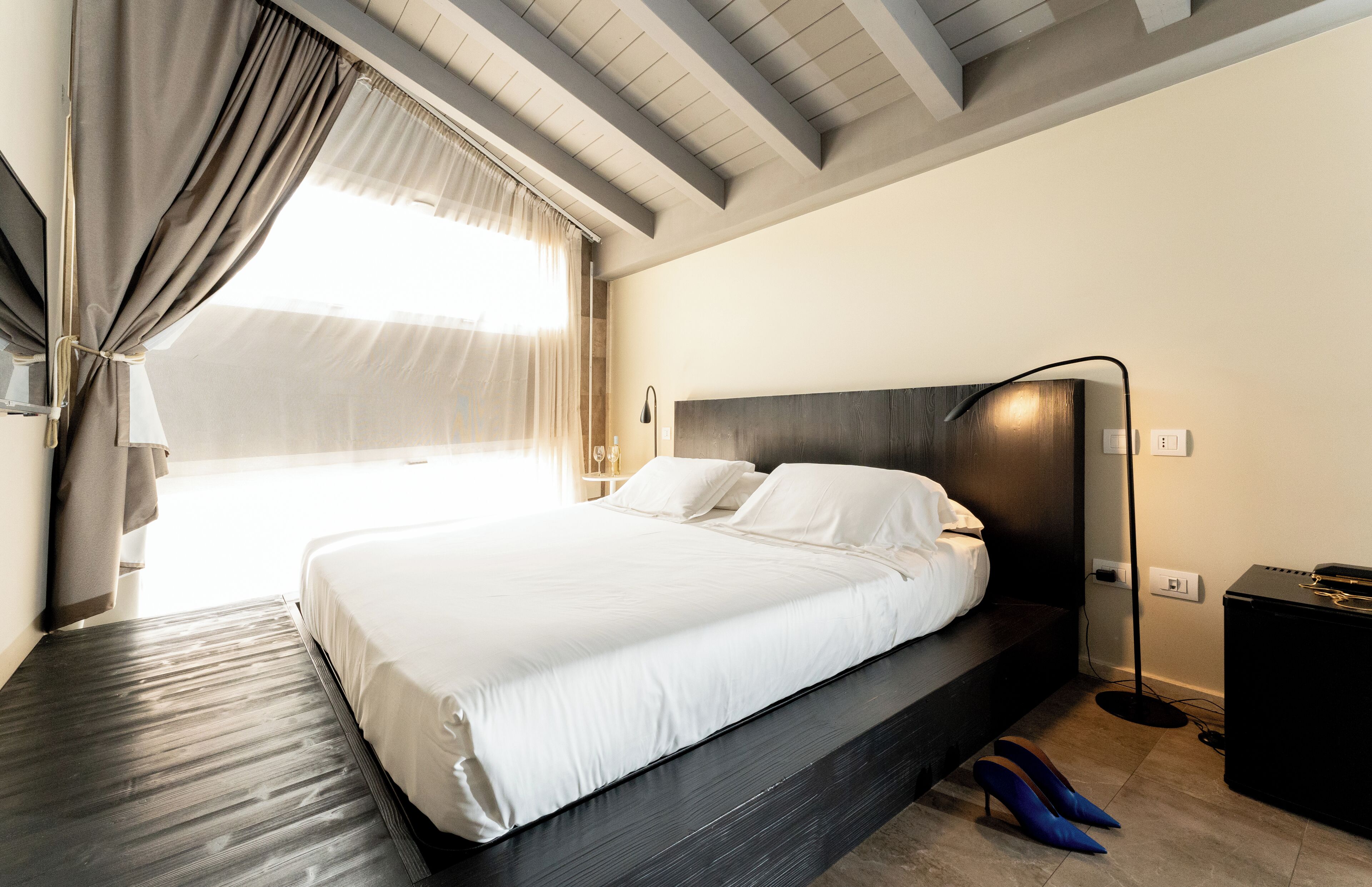Een moderne hotelkamer met een groot bed, een donker hoofdeinde, een nachtkastje, een lamp en uitzicht op het groen door dunne gordijnen.
