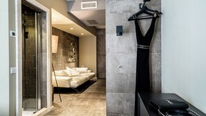 A modern hotel room with a glass shower, a beige sofa, a black dress on a hanger, and a black handbag on a table. - Neko Boutique Hotel (Cagliari)