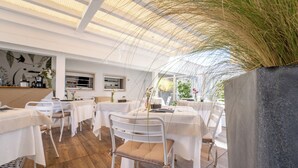 A restaurant with white tables and chairs, a wooden floor, and a large planter with grass. - Neko Boutique Hotel (Cagliari)
