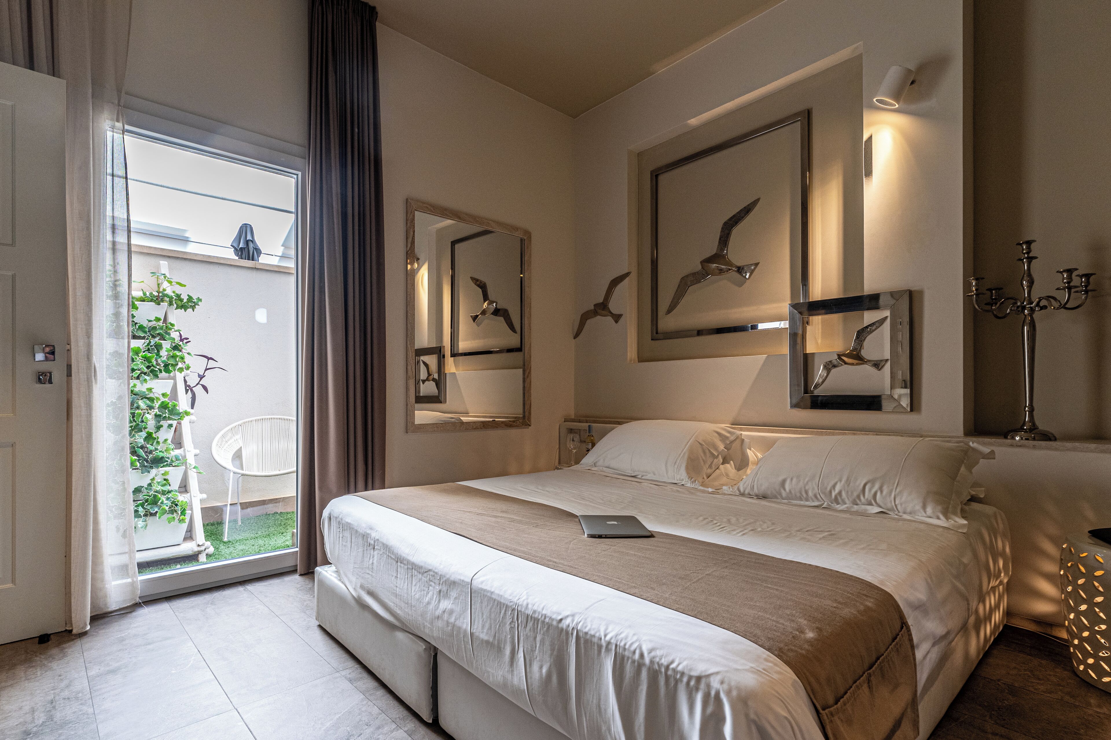 Une chambre avec un grand lit, un ordinateur portable posé sur le lit et une porte-fenêtre coulissante donnant sur un balcon agrémenté de plantes.
