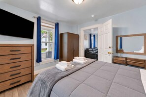 A hotel room with a bed, a wooden dresser, a wardrobe, a seating area with a chair, and a television mounted on the wall.