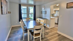 A dining area with a wooden table and white chairs, a kitchen in the background, and a clock on the wall. - Shortstop Apt 1 - All Star Baseball Rentals (Oneonta)