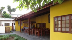 A yellow building with a covered patio area, wooden furniture, and a small garden.