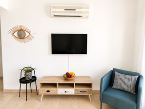 Un salon avec un téléviseur à écran plat fixé au mur, un meuble de rangement en bois en dessous, un fauteuil bleu, une petite table d’appoint avec une plante et un climatiseur mural.
