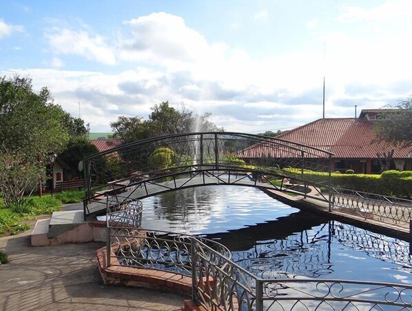 Un puente sobre una masa de agua que refleja el entorno, rodeado de vegetación y edificios con techos de tejas rojas.