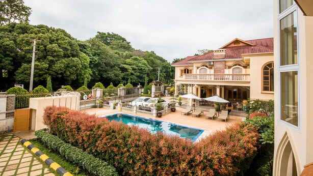 A pool area with lounge chairs and umbrellas, surrounded by lush greenery and a large house with a red roof. - Win Unity Pyin Oo Lwin (Pyin Oo Lwin)