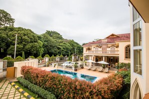 Un área de piscina con sillones de descanso y sombrillas, rodeada de exuberante vegetación y una casa grande con techo rojo.
