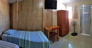 Una habitación con dos camas, un armario de madera, un escritorio con una silla, un ventilador de pie y un televisor montado en la pared.