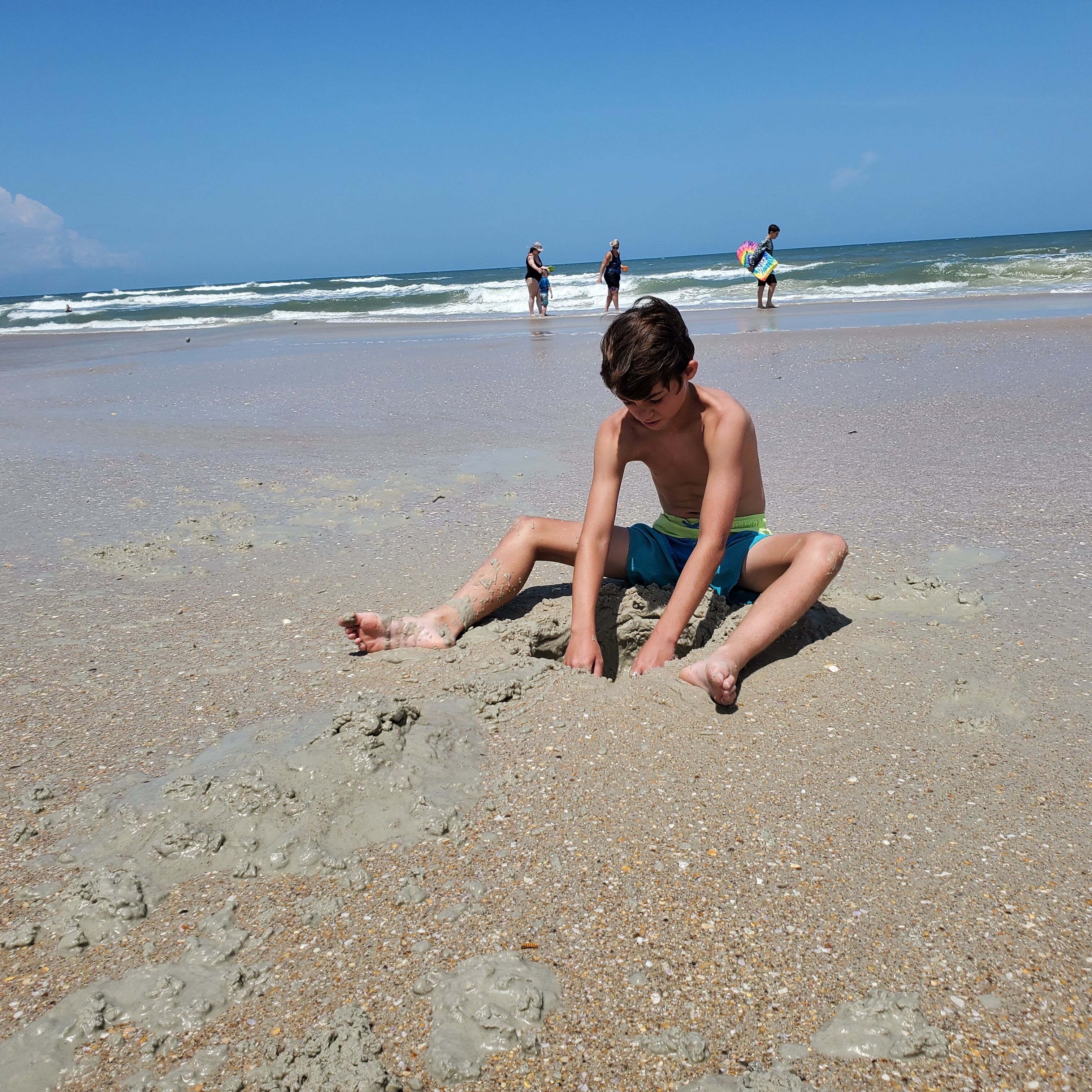 A beach scene with a child sitting on the sand, others in the background, and the ocean.